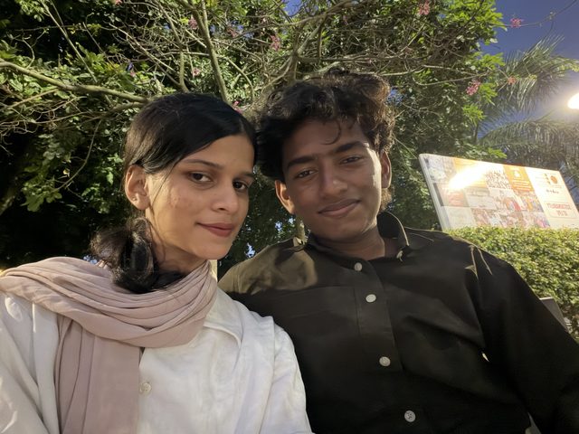Two friends sitting together outdoors beneath leafy branches.